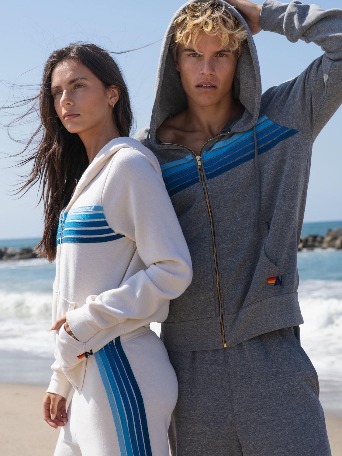 Two models wearing athletic tracksuits on a beach. The model on the left wears a white zip-up hoodie and matching pants with blue stripes, while the model on the right wears a heather gray zip-up hoodie and matching pants with blue diagonal stripes.